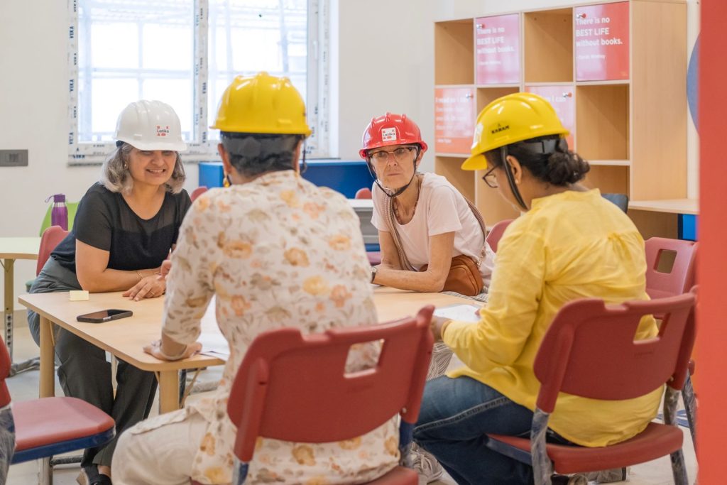 Four women, all wearing hard hats, sit around a children's table talking