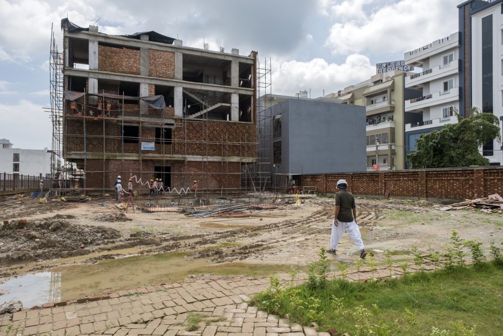 Man in dhoti and shirt walks across a muddly dirt lot towards a large brick building under construction.