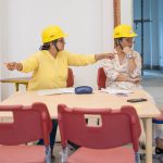 Two women, both in yellow hard hats, sitting at a child's table, having a heated conversation