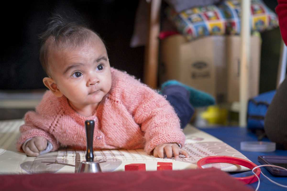 Baby in pink cardigan propped on her elbows on a play mat with a silver bell in front