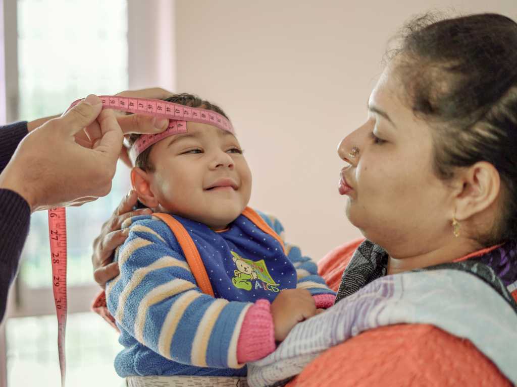 Woman measuring a small boy's head with a pink tape measure while another woman holds him