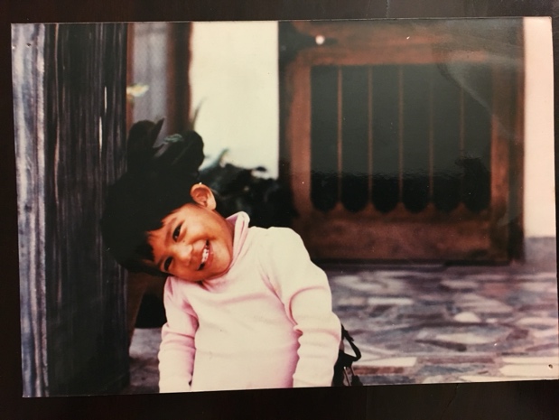 Young Moy Moy smiling warmly, leaning against a wall in a sunlit courtyard