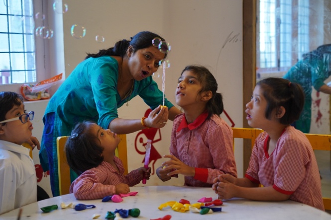 A Latika teacher blowing bubbles for three students in school uniforms at a classroom table