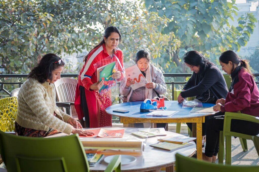 Latika mothers reading and sorting picture books together at outdoor tables on a sunny terrace