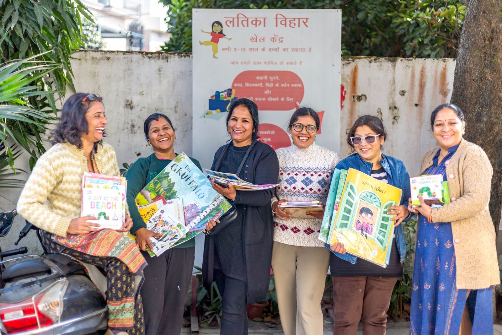 Six smiling Latika mothers holding armfuls of children’s books in front of the Latika Vihar sign