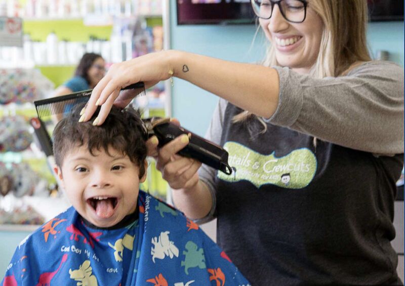 A delighted boy with Down syndrome getting a haircut from a beaming stylist at the Pigtails & Crewcuts salon