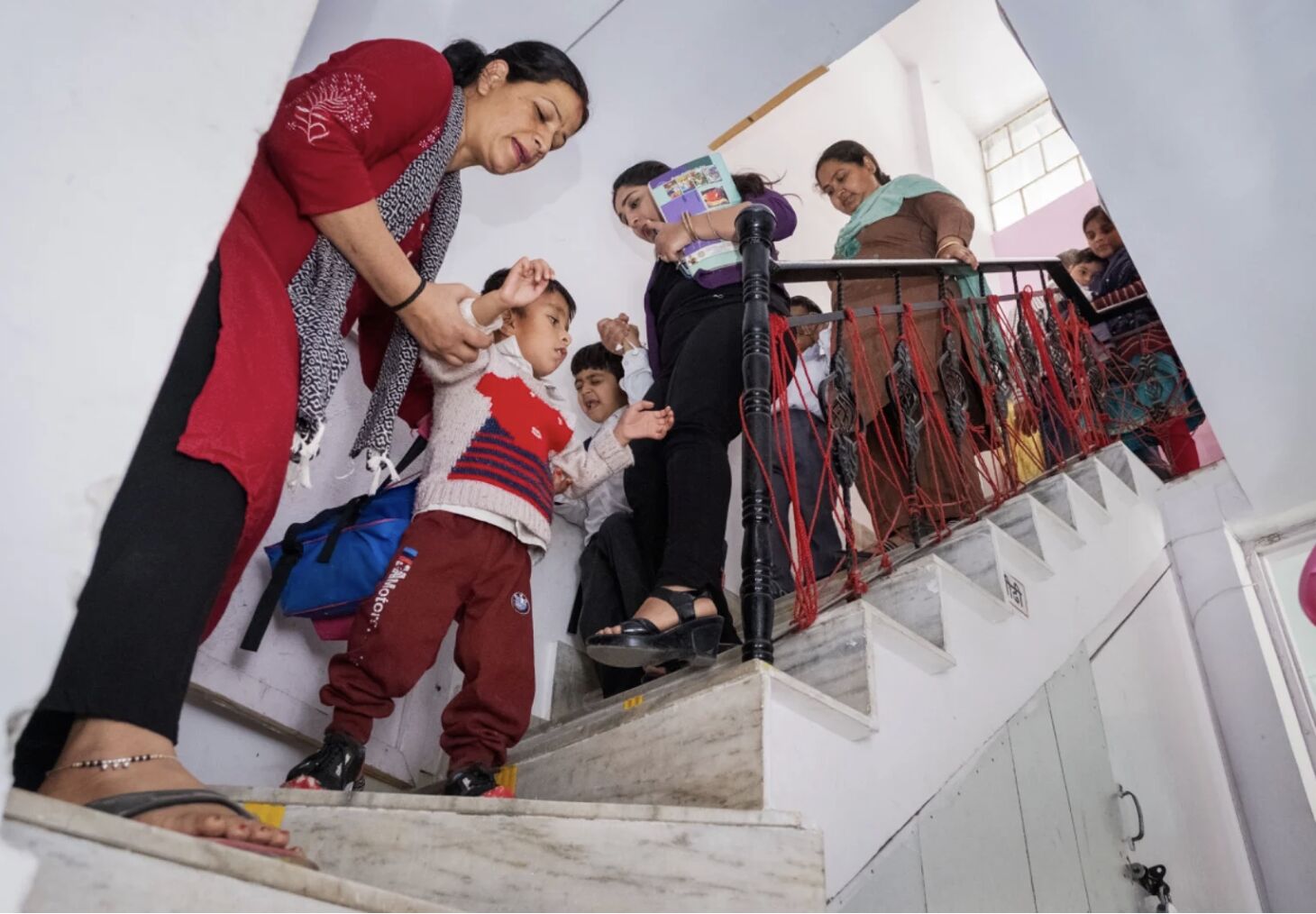 A Latika teacher in a red kurta helping a small student navigate down marble stairs, hand in hand