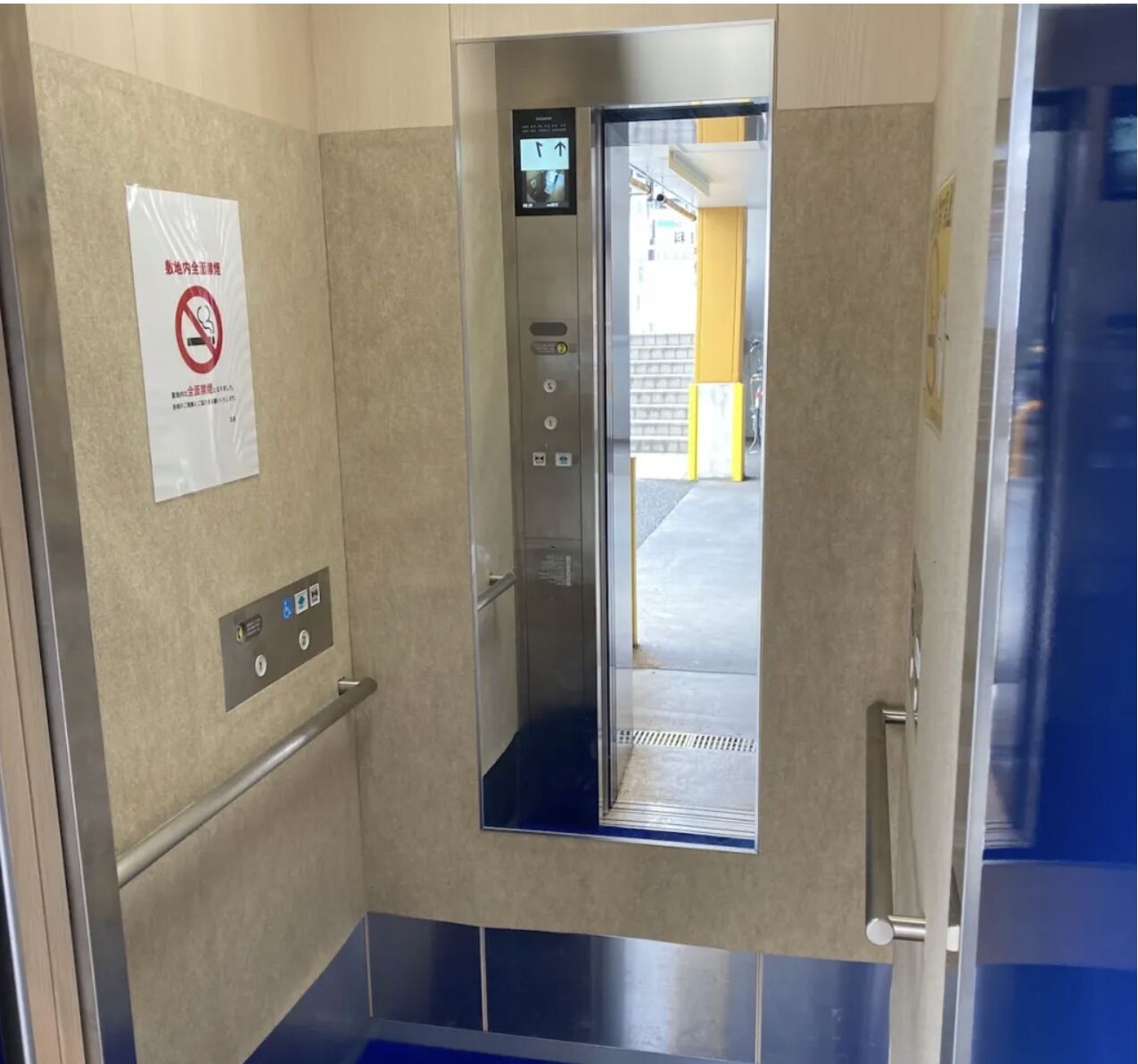 Inside a small accessible elevator with open doors and tactile controls, photographed as a reference for the new building’s lifts
