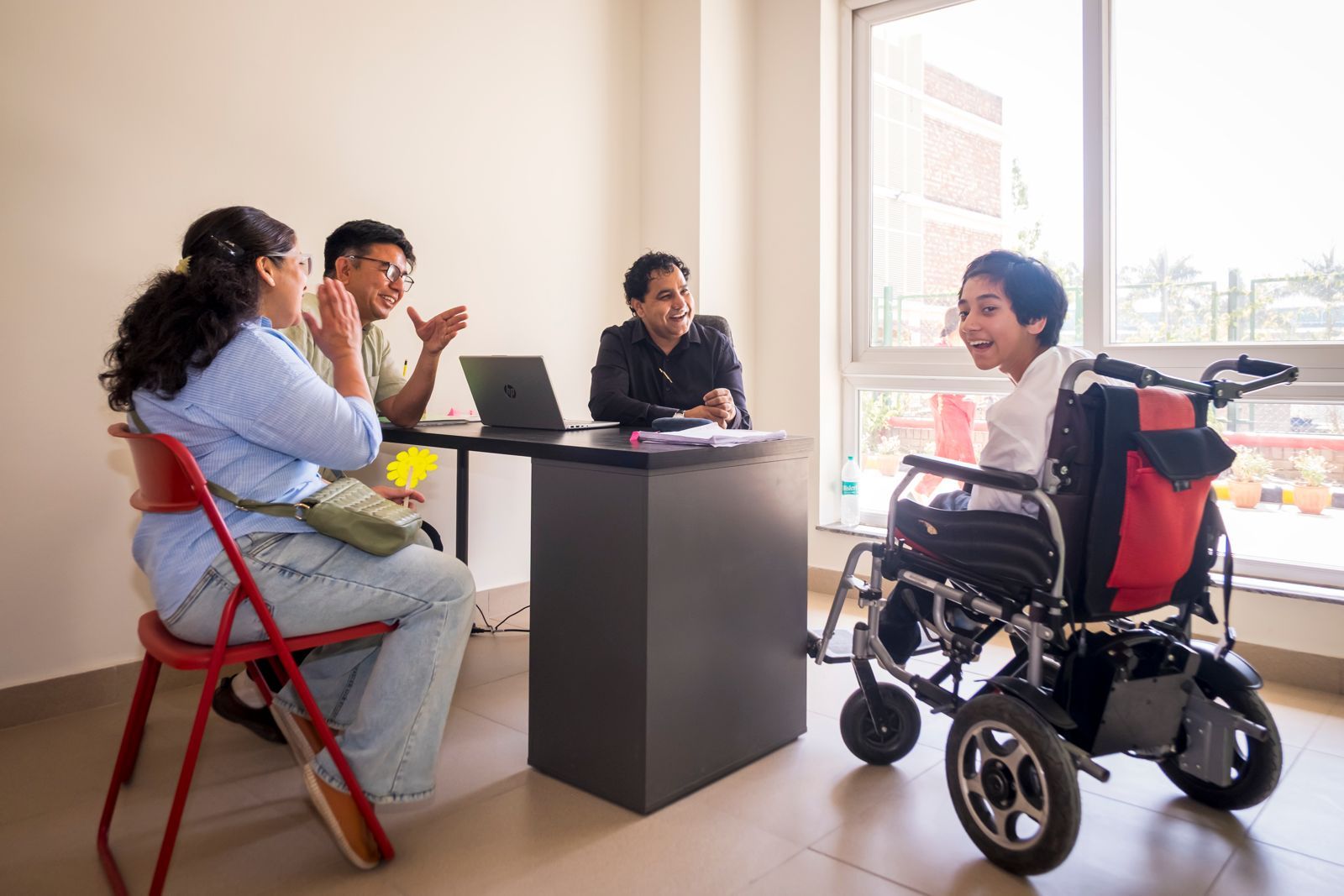 A Latika family meeting with two staff in the sunlit assessment room, a teenager in a motorised wheelchair joining the conversation