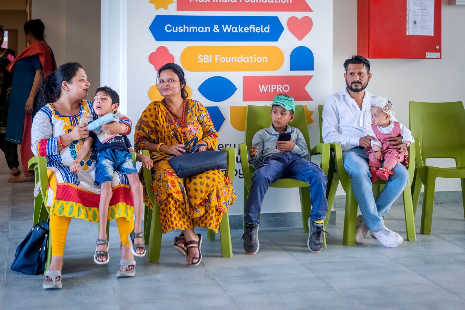 Families waiting on green chairs in front of a donor-recognition wall featuring Cushman & Wakefield, SBI Foundation, WIPRO and Max India Foundation