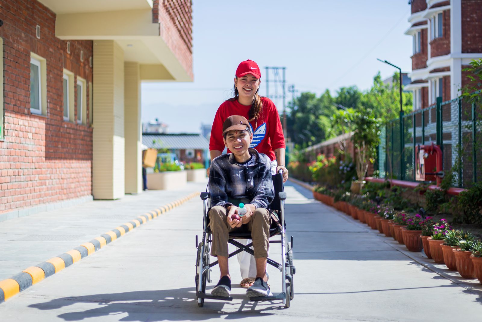A volunteer pushes a Latika student in a wheelchair along the bright outdoor walkway between buildings