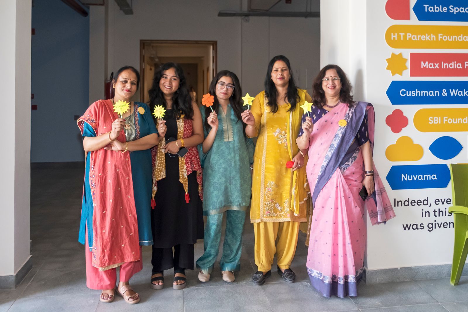 Five Latika friends in colourful Indian clothing posing with yellow paper stars in front of the donor wall