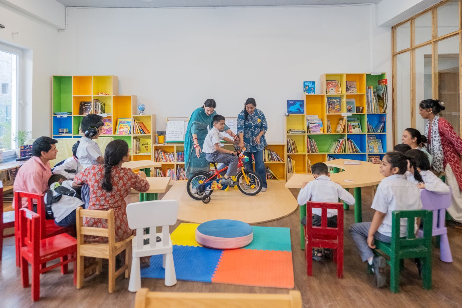 Children gathered around a small bicycle in the library while teachers help — colourful book displays in the background