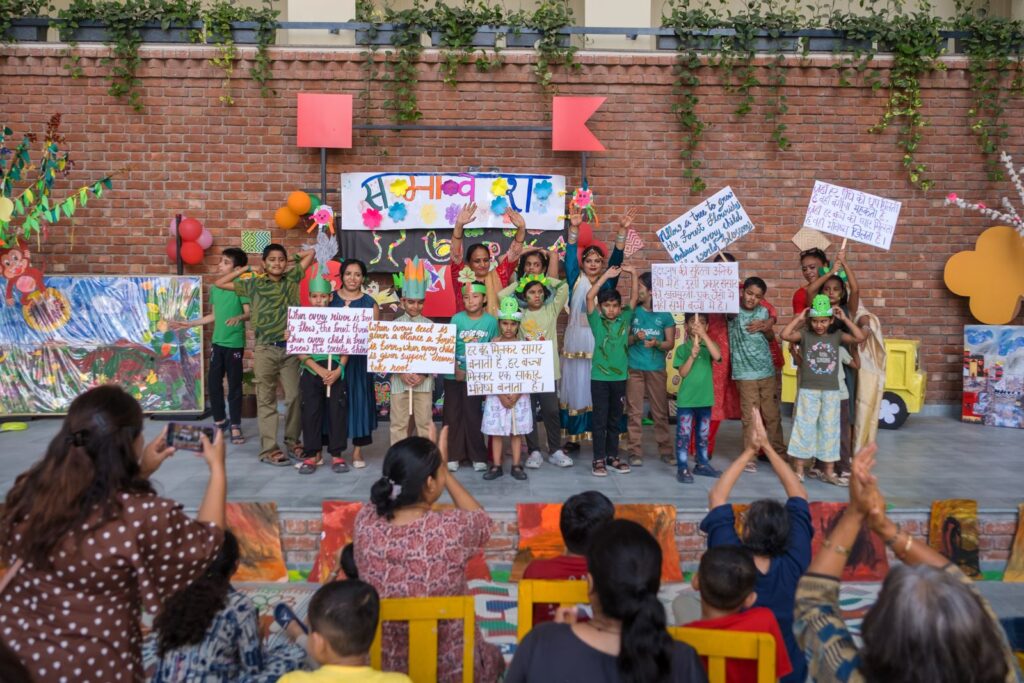 The full cast of children and teachers on stage at Samavesh 2026, hands raised, holding hand-painted placards with nature and inclusion poems