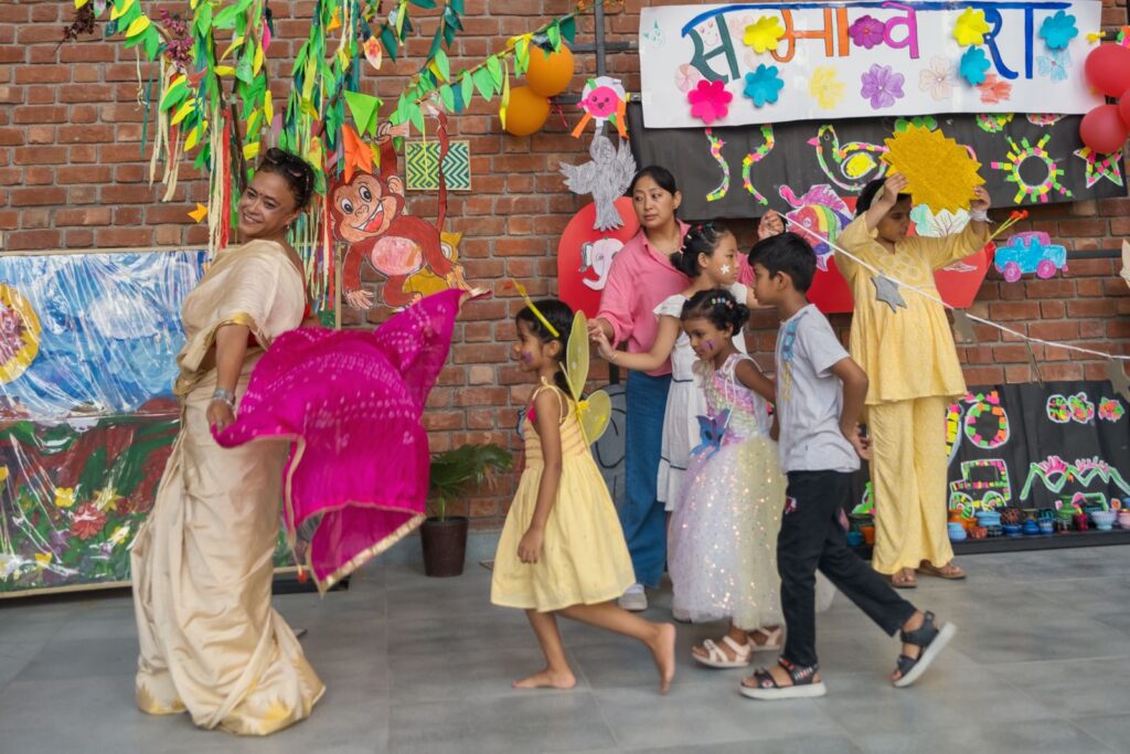 A teacher in a cream sari twirls as she leads a line of costumed children across the nature-themed Samavesh stage