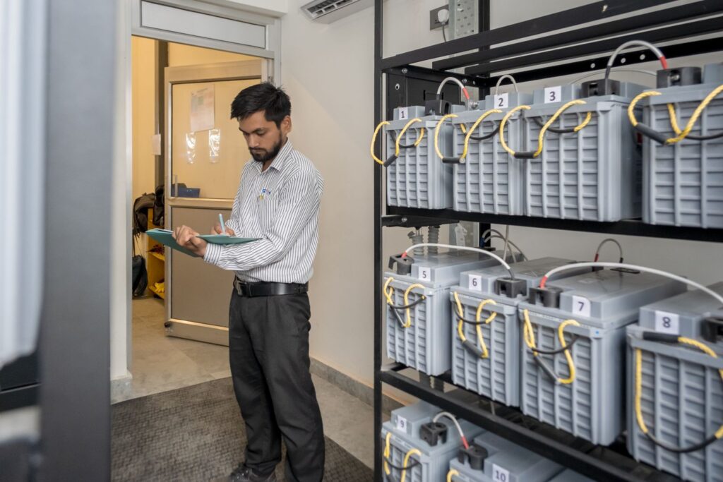 A staff member with a clipboard checking a rack of numbered backup batteries in the building's power room