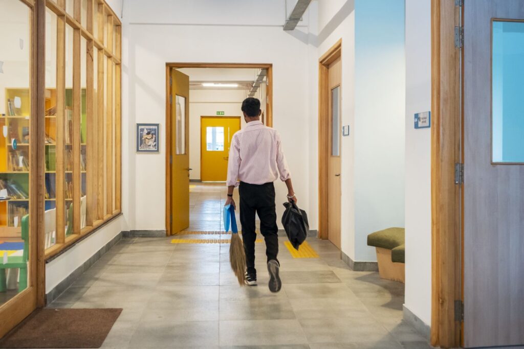 A staff member walking down a corridor carrying a broom and bag past the library, everyday upkeep in motion