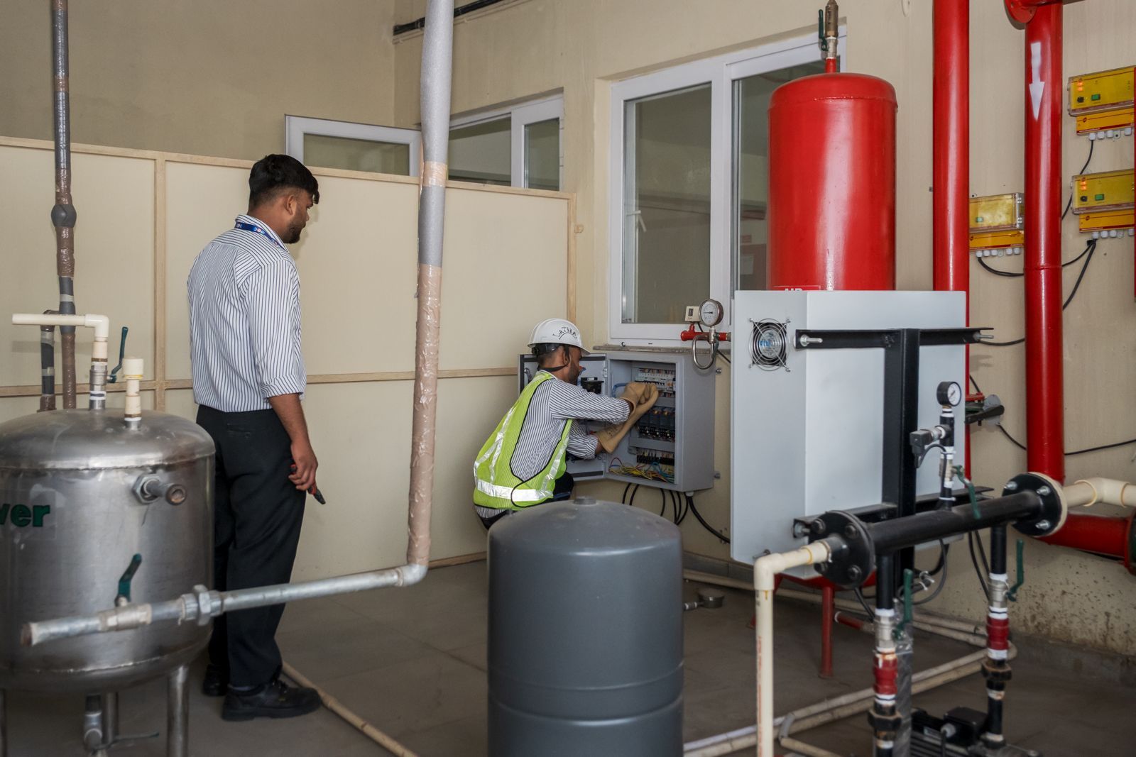 Two Latika staff service an electrical control panel in the building's plant room, beside water tanks, pipework and a red fire-suppression cylinder