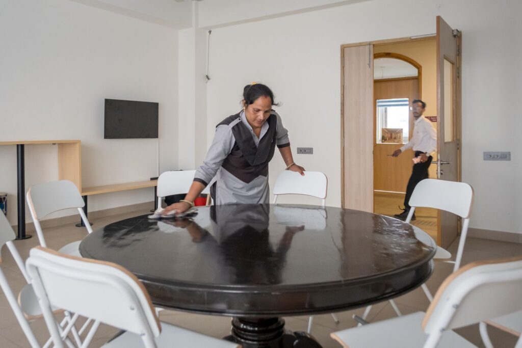 A housekeeping staff member polishing a round table in a tidy meeting room with white folding chairs