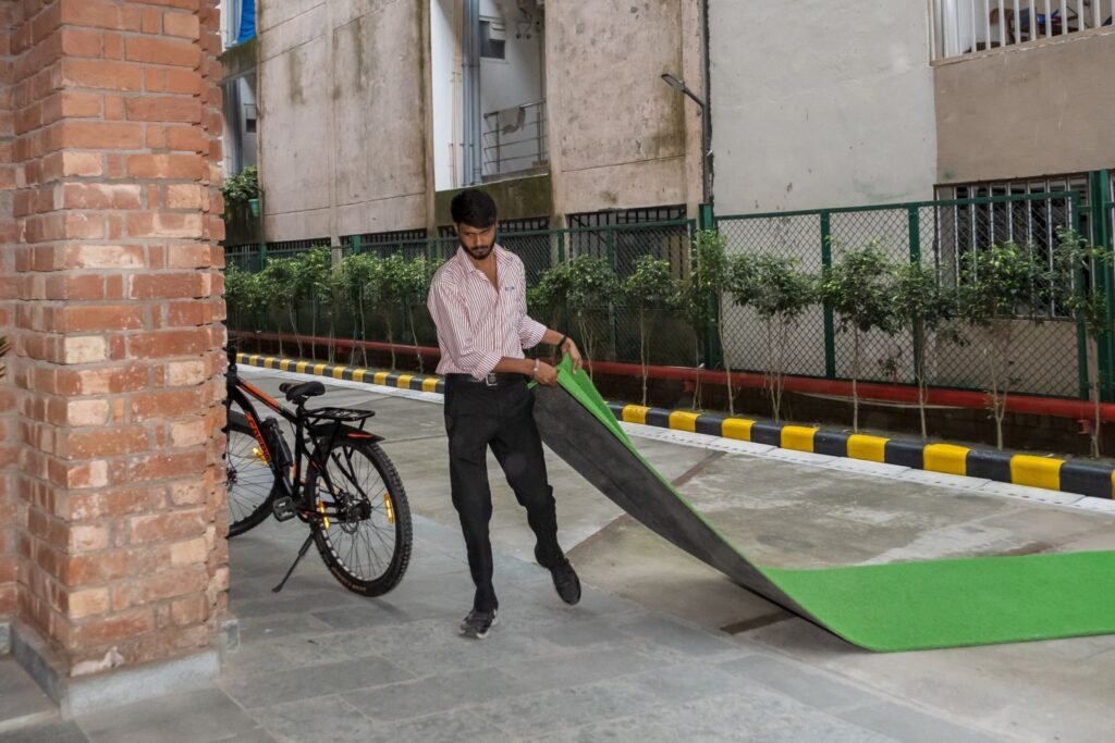 A young staff member unrolling a green mat along a paved outdoor walkway beside the building